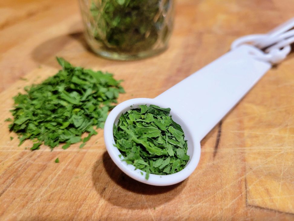 Measuring dried parsley in a tablespoon next to pile of dried parsley on a butcher block counter